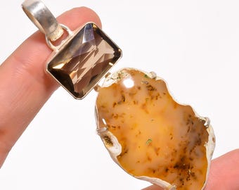 May include: A silver pendant featuring a large, irregularly shaped, light orange gemstone with brown inclusions. A rectangular, faceted, smoky quartz stone is set above it. The pendant is held by a hand.