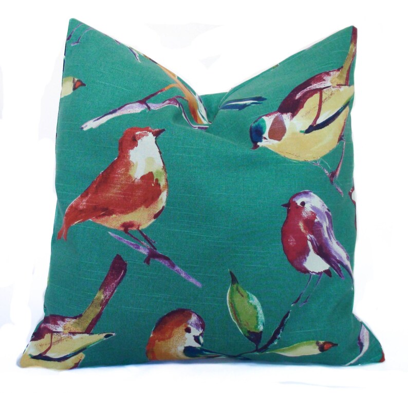May include: Teal pillow with a watercolor bird pattern. The birds are painted in shades of red, orange, yellow, blue, and purple. The pillow is made of a soft, textured fabric.