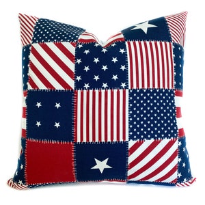 May include: Square decorative pillow with a patchwork design featuring red and white stripes, blue fields with white stars, and red squares. The pillow has a white background and is likely for home decor.