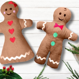 May include: Two gingerbread person plush toys, one with red hearts and a bow, the other with a green button and a red bow tie. The toys are on a white wooden background with a brown gift box wrapped with twine and a red holly tag.