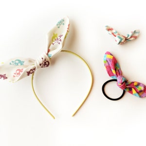 May include: Three fabric hair accessories. One headband with a white fabric bow with a colorful skull pattern. One hair tie with a pink, blue, and yellow fabric bow. One hair tie with a white and blue fabric bow.