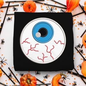 May include: A black t-shirt with a white and red embroidered eyeball design. The eyeball is blue with a black pupil and a white highlight. The t-shirt is surrounded by orange pumpkins and black spider webs.