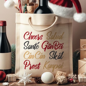 May include: A white canvas tote bag with a red Santa hat on top. The bag has the words "Cheers, Salud, Santé, Gan Bei, Cin Cin, Skål, Prost, Kanpai" printed on it in different colors. The bag is surrounded by Christmas decorations, including a bottle of wine, a roll of wrapping paper, and a few ornaments.