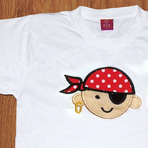 Op de afbeelding: Wit T-shirt met een geborduurd cartoon-piratengezicht dat een rood-wit geblokt bandana en een ooglapje draagt. De piraat heeft een gouden oorring.