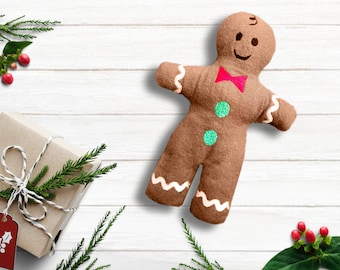 May include: A brown gingerbread man plush toy with a red bow tie and green buttons. The toy is lying on a white wooden surface with green sprigs and red berries.