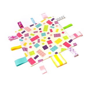 Puede incluir: Una manta blanca para bebé con un patrón de bloques de Lego coloridos y múltiples etiquetas de cinta de tela en rosa, amarillo, azul y verde.