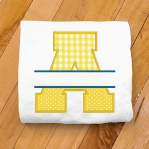 May include: A white t-shirt with a yellow and white gingham letter A and a blue line above and below the letter. The letter A is outlined in yellow.