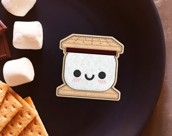 May include: A brown and white felt s'more patch with a smiling marshmallow face. The patch is surrounded by graham crackers, chocolate squares, and marshmallows on a black plate.