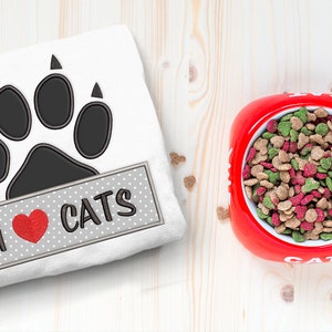 May include: A white t-shirt with a black paw print and a gray banner that says "I love cats" with a red heart. A red cat food bowl with cat food inside is on a wooden surface.