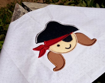 May include: A white fabric with an embroidered design of a cartoon pirate with a black hat, red bandana, and brown pigtails.