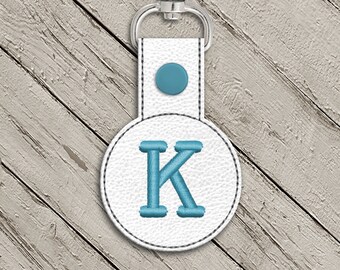 May include: White leather keychain with a blue embroidered letter K on a round tag. The keychain has a silver metal ring.