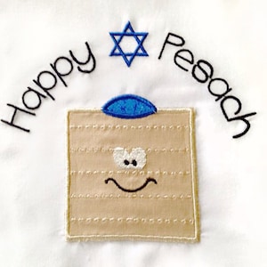 Può includere: Tessuto bianco con il testo ricamato "Happy Pesach" e una stella di David. Un matzà di cartone animato con una kippah blu e un viso sorridente è ricamato sotto il testo.