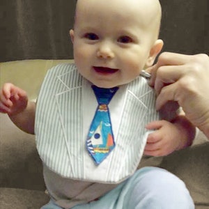 May include: A baby wearing a white bib with a blue and white striped pattern and a blue tie with a sailboat design. The bib has a white collar and a white button closure.