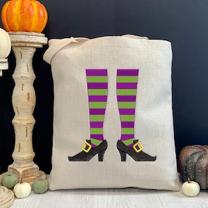 May include: A white canvas tote bag with a green and purple witch's legs and black shoes design. The bag has a long handle and is perfect for carrying everyday essentials.