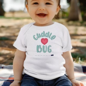 May include: A white baby bodysuit with the words "Cuddle Bug" in teal script and a pink heart. The baby is wearing blue jeans and is sitting on a blanket outdoors.