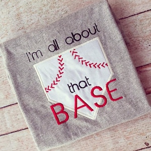May include: Gray t-shirt with a white baseball home plate design and red stitching. The text "I'm all about that BASE" is embroidered on the shirt.