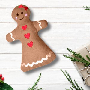 Puede incluir: Un juguete de peluche de jengibre marrón con corazones rojos y ribetes blancos sobre un fondo de madera blanca. El juguete está al lado de un regalo envuelto con una etiqueta roja.