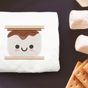 May include: A white t-shirt featuring an embroidered image of a smiling marshmallow with chocolate and graham cracker, resembling a s'more.