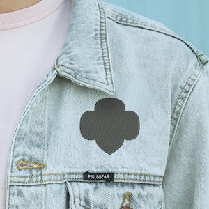 May include: A light blue denim jacket with a dark brown patch in the shape of a four-leaf clover. The jacket has a label that says "PULL&BEAR".