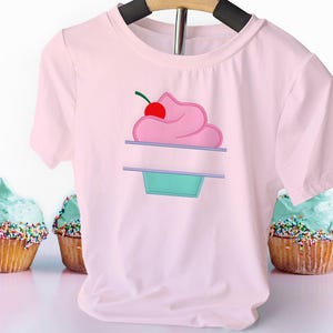 May include: A pale pink t-shirt with a cupcake design. The cupcake has pink icing, a red cherry, and a teal base. The shirt is hanging on a wooden hanger, with cupcakes in the background.