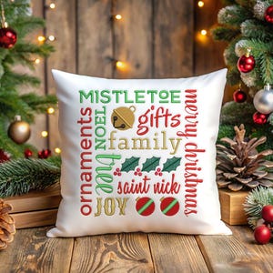 May include: White square pillow with Christmas-themed text in red, green, and gold. The text includes words like "Mistletoe", "Gifts", "Family", and "Merry Christmas". The pillow is on a wooden surface with Christmas decorations in the background.