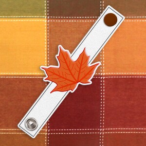 May include: A white fabric strap with a brown button and an orange maple leaf design. The strap is on a plaid background with orange, brown, and green squares.