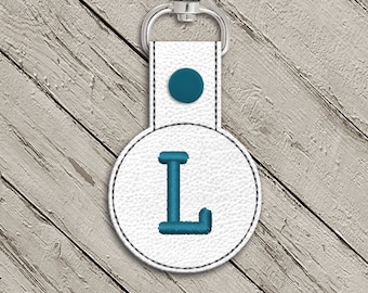 May include: White leather keychain with a teal blue circle and the letter L embroidered in teal blue.