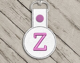 May include: A white leather keychain with a pink letter Z embroidered on a round tag. The keychain has a pink button and a silver metal ring.