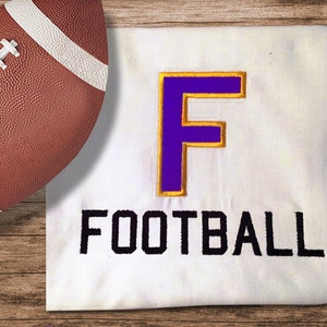 Puede incluir: Una camiseta blanca con una gran "F" morada bordeada de oro y la palabra "FOOTBALL" en letras negras. Un balón de fútbol marrón está en el fondo.