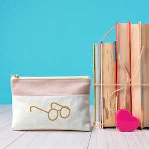May include: A white and pink pencil case with a gold embroidered glasses design. A stack of books tied with twine and a pink heart-shaped object are to the right. The background is a solid teal color.