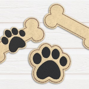 Könnte beinhalten: Drei Filzpatches mit Hundeknochen- und Pfotenabdruck-Designs. Die Knochenpatches sind beige mit braunen Nähten. Die Pfotenabdruckpatches sind schwarz mit braunen Nähten.