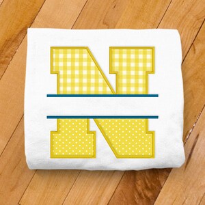 以下が含まれることがあります： 白地に黄色のNの文字が刺繍されたTシャツ。Nの文字はギンガムチェックと水玉模様で、中央に青い線が引かれている。