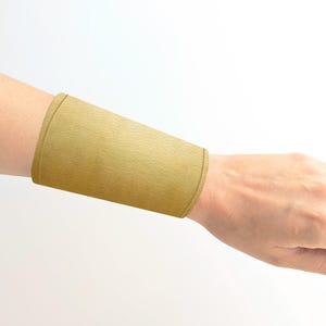 Puede incluir: Un brazalete de tela de color dorado. El brazalete está hecho de un material suave y texturizado y está diseñado para usarse en el antebrazo.