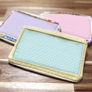 May include: Three rectangular fabric coasters with gingham patterns. One is light blue and white, another is pink and white, and the third is purple and white. Each coaster has a patterned border.