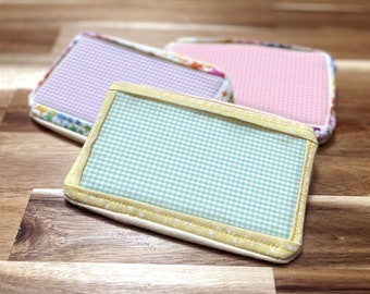 May include: Three rectangular fabric coasters with gingham patterns. One is light blue and white, another is pink and white, and the third is purple and white. Each coaster has a patterned border.