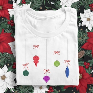 May include: White t-shirt with a design of five colorful Christmas ornaments hanging from red bows.