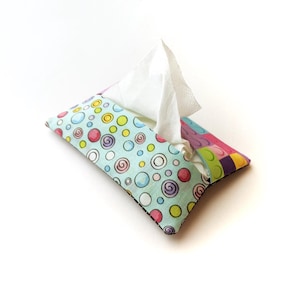 May include: A fabric tissue box cover with a whimsical design. The cover features a light blue background with colorful circles and swirls. The right side has a patchwork design. White tissues are visible.