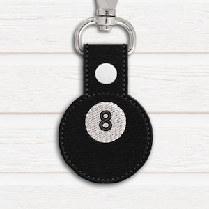 Puede incluir: Llavero de cuero negro con un diseño de bola 8 blanca. El llavero tiene un anillo de metal plateado.