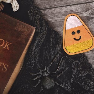 May include: A Halloween still life featuring a brown book titled "Book of Secrets" with gold lettering, a candy corn patch with a smiling face, a black spider, and black fabric on a wooden surface.