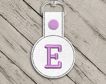 May include: White leather keychain with a round, white circle featuring a pink embroidered letter E. The keychain has a silver metal ring.