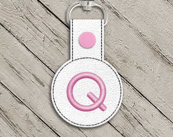 May include: White leather keychain with a pink circle and the letter Q embroidered in pink.