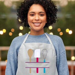 May include: A light grey and white striped apron featuring wooden spoon, spatula, and whisk illustrations. The apron has a purple horizontal line and red, pink, and blue accents. The person in the image is wearing a blue jumper.