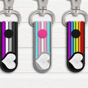Może przedstawiać: Trzy breloki z różnymi wzorami flag LGBT. Każdy brelok ma czarno-białe serce z kółkiem w środku. Pierwszy brelok jest czarny, biały, fioletowy i szary. Drugi brelok jest jasnoniebieski, różowy i biały. Trzeci brelok jest tęczowy.