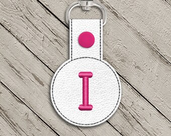 May include: White leather keychain with a pink circle button and a pink embroidered letter I on a white circle.