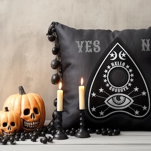 May include: A Halloween display with carved pumpkin decorations, lit candles, and a black Ouija board. A black cushion with the words "YES" and "NO" is in the background. The pumpkins have carved skull faces.