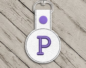May include: White leather keychain with a purple letter P embroidered on a round circle. The keychain has a silver metal ring and a purple button.