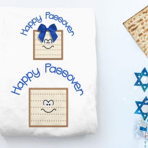May include: White fabric with blue embroidery that says "Happy Passover" and features two images of matzah with a smiling face.  A blue bow is on top of the matzah in the top image.