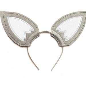 May include: A grey and white wolf ear headband. The ears are made of grey felt with white inner ear details and a silver wire headband. The ears have a pointed shape and are outlined in a darker grey.