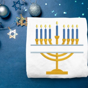 May include: A white t-shirt with a gold embroidered menorah with nine blue candles. The menorah is on a blue background with silver glitter and silver stars.