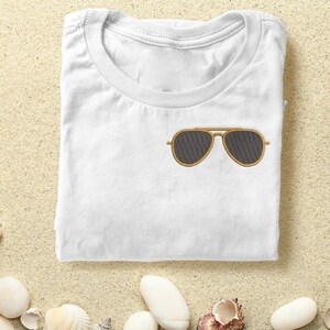 May include: White t-shirt with a pair of brown aviator sunglasses embroidered on the front. The t-shirt is folded and lying on a sandy surface.